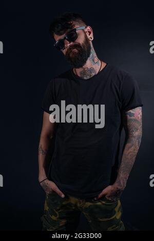 Bärtiger Mann Modell mit Tattoos und Hände in der Tasche, gekleidet in einem schwarzen T-Shirt und Sonnenbrillen Posen auf schwarzem Hintergrund. Stockfoto