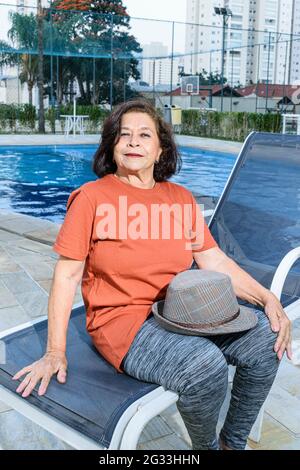 Eine lächelnde 74-jährige Frau, die am Rand des Pools mit einem Hut auf ihrem Schoß sitzt. Stockfoto