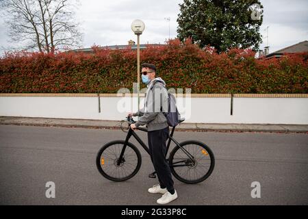 Ein junger Mann drückt mit den Händen auf sein Fahrrad, steht auf einer Straße und trägt eine OP-Maske, um während der Coronavirus-Pandemie sicher zu bleiben. Stockfoto