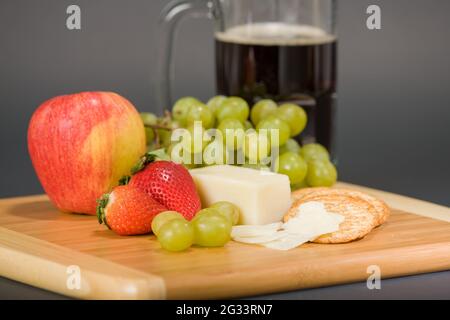 Gruyere Schweizer Käse, Becher Bier, Obst und Vollkorncracker auf einem hölzernen Schneidebrett Stockfoto