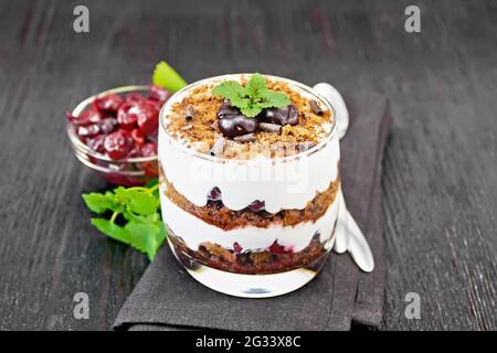 Dessert Schwarzwald mit Kirschen, Schokoladenkeks und weichem Quark mit Creme im Glas auf Serviette, Minze, Beeren in einer Schüssel und einem Löffel auf BL Stockfoto