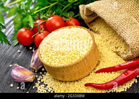 Roher Couscous in einer Schüssel und ein Sack mit Sackleinen, Knoblauch, Tomaten, Paprika und Kräutern auf Holzbrett Hintergrund Stockfoto