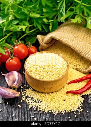 Roher Couscous in einer Schüssel und ein Sack mit Sackleinen, Knoblauch, Tomaten, Paprika und Kräutern auf dem Hintergrund eines dunklen Holzbretts Stockfoto
