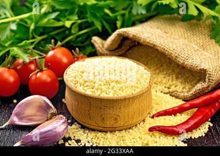 Roher Couscous in einer Schüssel und ein Sack mit Sackleinen, Knoblauch, Tomaten, Paprika und Kräutern auf schwarzem Holzbrett Hintergrund Stockfoto
