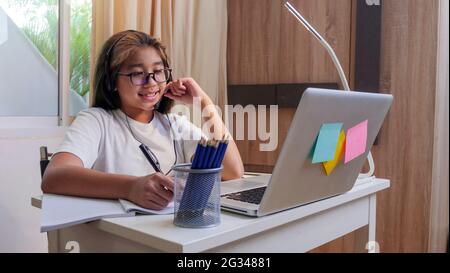 Asiatische Mädchen in Kopfhörer sitzen am Schreibtisch Studie online auf Laptop. Kinder tragen Headset-Handschrift beim Lernen im Notebook mit Hilfe von Internet-Lektionen über Quarantäne. G Stockfoto