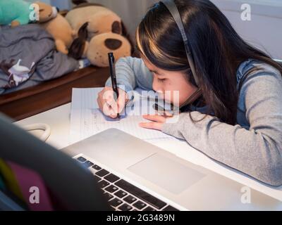 Asiatische Mädchen in Kopfhörer sitzen am Schreibtisch Studie online auf Laptop. Kinder tragen Headset-Handschrift beim Lernen im Notebook mit Hilfe von Internet-Lektionen über Quarantäne. G Stockfoto