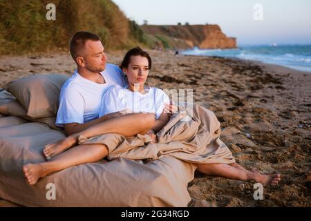 Junges Paar eingewickelt in eine Decke im Freien. Glückliches Paar in der Liebe liegen und rotieren im Bett am Meer bei Sonnenuntergang. Glückliches kaukasisches Ehepaar von Ehemann und Ehefrau, eingewickelt in eine Decke im Freien Stockfoto
