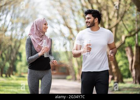 Glückliches arabisches Paar in aktiver Form, das im Sommerpark mit einer Flasche Wasser in den Händen zusammenläuft. Junge Männer und Frauen verbringen ihre Freizeit aktiv an der frischen Luft. Stockfoto