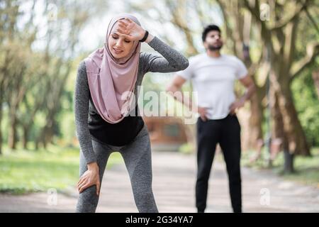Erschöpfte Frau in Sportkleidung und Hijab, die beim Training im Freien Pause einnahm. Hübscher muslimischer Kerl, der sich auf dem Hintergrund ausruht. Konzept des aktiven Lebensstils. Stockfoto