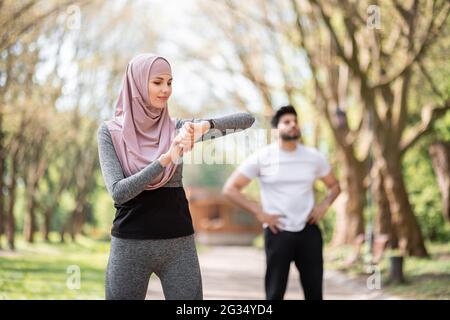 Junge Frau in Hijab Überprüfung Zeit auf Smart Watch, während muslimische Kerl entspannen nach dem Training auf Hintergrund. Gesundes Sportpaar trainiert gemeinsam im Park. Stockfoto