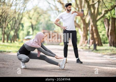 Aktive junge Frau im Hijab, die flexible Übungen macht, während ein hübscher arabischer Mann hinter sich steht und eine intelligente Uhr anschaut. Konzept von Menschen, Technologie und Aktivität. Stockfoto
