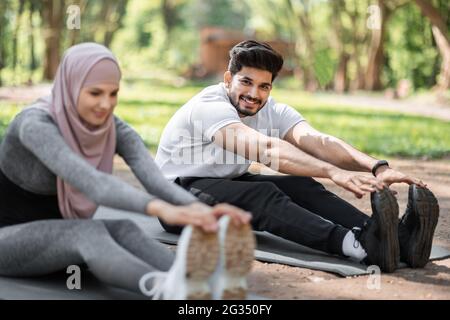 Arabischer Mann und Frau im Hijab wärmen sich vor dem Training im Freien auf einer Yogamatte auf. Glückliche junge Familie, die einen gesunden und aktiven Lebensstil führt. Sportkonzept. Stockfoto