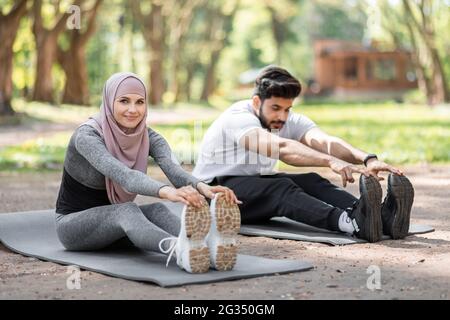 Aktives muslimisches Paar in Sportkleidung auf Yogamatte sitzend und Beine streckend. Junge Frau im Hijab und bärtiger Mann trainieren gemeinsam an frischer Luft. Stockfoto