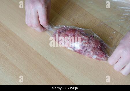 Schritt für Schritt Zubereitung von hausgemachtem gehacktem Schinken. Schritt 10: Die Hände eines Mannes drehen eine Portion mariniertes Fleisch in eine Rolle. Roher Schinken in Lebensmittelverpackung. C Stockfoto