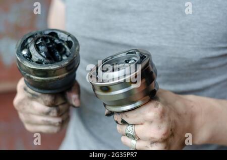 Ein junger Erwachsener hält ein neues und altes Antriebswellengelenk in der Hand. Ein Mann mit silbernen Ringen an den Fingern, der Autoteile hält. Auto-Service. Stockfoto