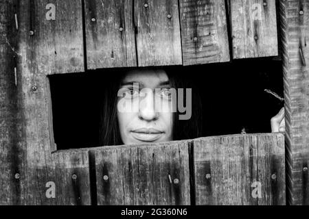 Junge Frau schaut aus nächster Nähe durch den Riss eines verschlossenen Holzschuppens. Schwarzweiß-Foto. Stockfoto