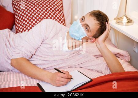 Ein junger Mann mit europäischem Auftritt in lässiger Kleidung in einer Schutzmaske, mit einem nachdenklichen und verträumten Gesichtsausdruck, liegt mit einem Notizbuch und schreibt mit einem Stift darin Stockfoto