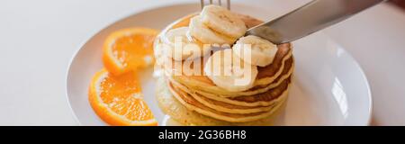 Nahaufnahme der Messer schneiden Pfannkuchen mit Orangen-und Bananenscheiben auf weißem Teller, Banner Stockfoto