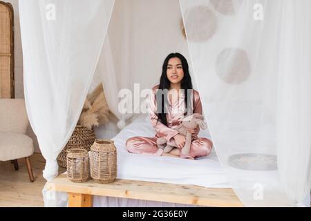 Junge asiatische Frau im Seiden-Schlafanzug sitzt mit gekreuzten Beinen auf dem Bett und hält weiches Spielzeug im Schlafzimmer Stockfoto