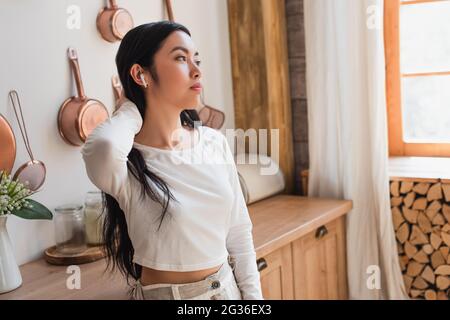 Müde junge asiatische Frau in weißer Bluse, die mit Ohrhörern steht und die Hand in der Küche am Hals hält Stockfoto