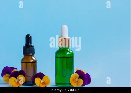 Serum-Glasflaschen mit Pipette und schönen Viola-Blüten auf blauem Hintergrund. Natürliches organisches Spa-Kosmetikkonzept. Vorderansicht Stockfoto