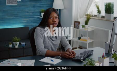 Porträt einer fröhlichen schwarzen Frau, die während der Heimsuchung am Schreibtisch im Wohnzimmer auf die Kamera schaut. Lächelnder Schüler, studiert am Computer in den Nachrichten der High School Stockfoto