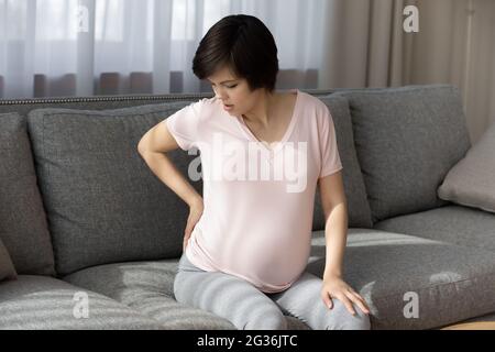 Müde, schwanger, die den Rücken berührt, massiert, auf der Couch sitzend Stockfoto