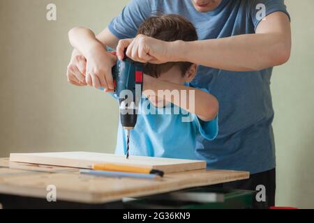 Vater und Sohn arbeiten zusammen, während sie an der Werkbank stehen. Kleiner Junge lernt zu bohren. Nettes Kind hält Bohrschrauber und Mann hilft ihm Stockfoto