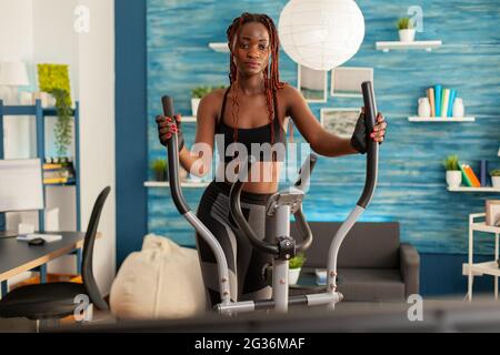 Black Person Training im Wohnzimmer mit einem Crosstrainer für das Cardio-Training und Online-Training. Sportlich starke aktive Frau mit gesundem Lebensstil. Stockfoto