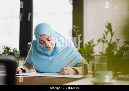 Lächelnder arabischer Ingenieur in Hijab zeichnet Blaupause auf verschwommenem Vordergrund Stockfoto