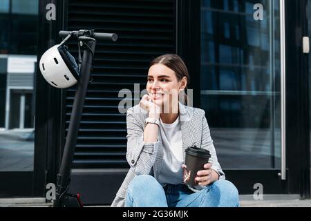 Schöne lächelnde Geschäftsfrau mit Kaffee sitzt auf einem Elektroroller auf Gebäude wegschauen Stockfoto