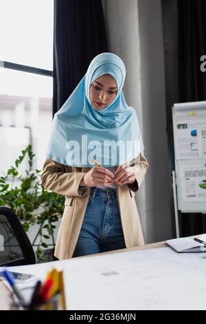 Junger muslimischer Ingenieur, der den Stift hält, während er im Büro die Blaupause ansieht Stockfoto