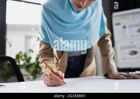 Beschnittene Ansicht eines verschwommenen muslimischen Ingenieurs, der im Büro Blaupause zeichnete Stockfoto