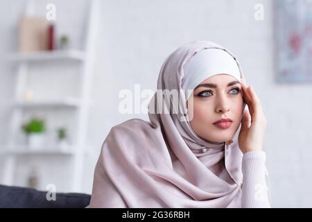 Nachdenkliche muslimische Frau im Hijab, die Hand in der Nähe des Gesichts hält, während sie zu Hause wegschaut Stockfoto
