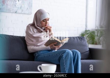 Hübsche muslimische Frau, die das Buch auf dem Sofa liest, fast eine verschwommene Tasse Tee Stockfoto