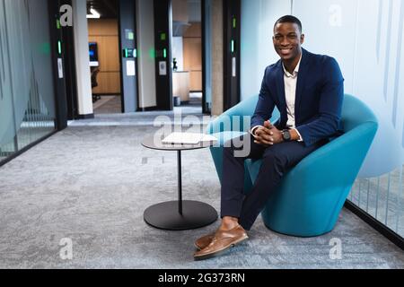 Porträt eines afroamerikanischen Geschäftsmannes, der lächelt, während er auf einem Stuhl im modernen Büro sitzt Stockfoto