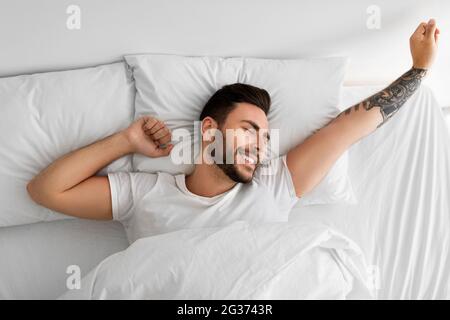 Konzept des Erwachens. Glücklich ausgeruhter, gut gelaunter Mann, der morgens aufwachte und sich die Hände streckte, im Bett lag, Draufsicht Stockfoto