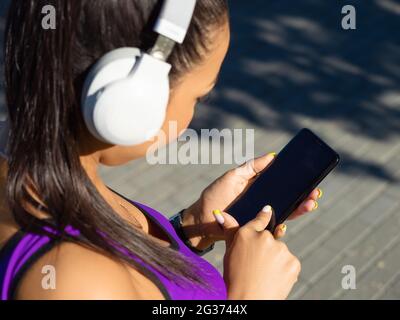 Junge asiatische Mädchen in weißen Kopfhörern wählt Musik zum Joggen in einer mobilen Anwendung. Das Konzept moderner Technologien im Sport. Stockfoto