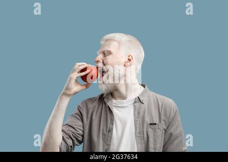 Essen Sie Früchte. Junger Albino-Mann beißt frischen roten Apfel und steht auf türkisfarbenem Studiohintergrund Stockfoto