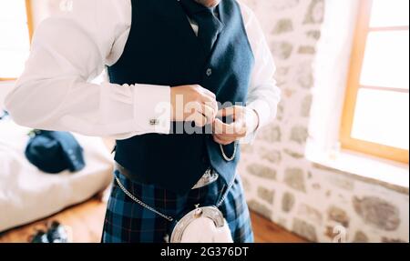 Ein Mann in schottischem Nationalkleid knöpft seine Weste hoch und bereitet sich in Nahaufnahme auf eine Hochzeitszeremonie in einem Hotelzimmer vor Stockfoto