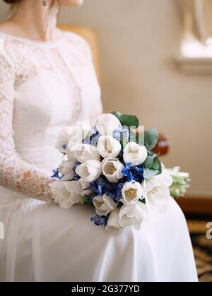 Die Braut im Hochzeitskleid sitzt und hält in ihren Händen einen zarten Strauß mit weißen und blauen Blumen Stockfoto