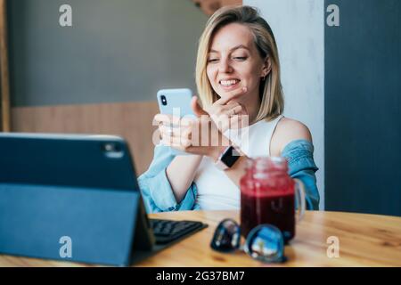 Junge fröhliche, fröhliche Blondine arbeitet auf einem Laptop und Handy und sitzt an einem Tisch in einem Café Stockfoto