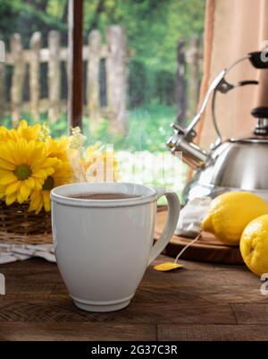 Tasse heißen Tee mit Zitronen und Blumen auf einem Holztisch an einem Fenster mit ländlichem Hintergrund Stockfoto