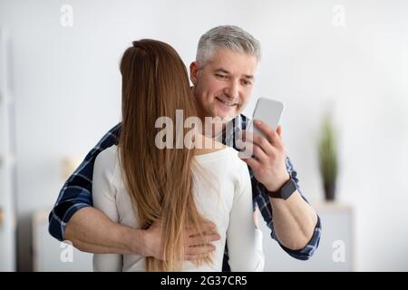 Reifer Mann umarmt seine Frau, steckt im Smartphone fest, achtet nicht, schreibt seinem Geliebten eine SMS, durchläuft eine Familienkrise Stockfoto