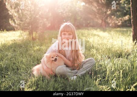 Nettes lächelndes Kind Mädchen 6-7 Jahre alt mit Spaß mit Haustierhund sitzen im Park auf grünem Rasen im Freien über Sonnenlicht. Sommersaison. Freund Stockfoto