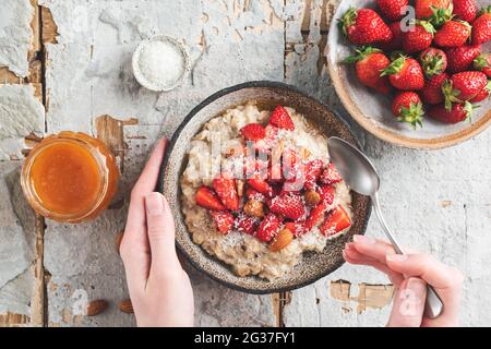 Haferflockenschale mit Erdbeeren, Mandeln und Kokosnuss in weiblichen Händen. Diät, sauberes Essen, Schlankheitskonzept. Draufsicht Stockfoto