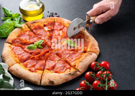 Hand schneiden Pepperoni Pizza auf schwarzem Beton Hintergrund. Leckere heiße italienische Pizza in Scheiben geschnitten Stockfoto