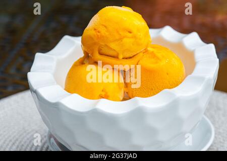 Drei Kugeln gelbes Eis in einer weißen Schüssel auf dem Tisch im Sommercafé. Eisiger Nachtisch mit Mangogeschmack Stockfoto