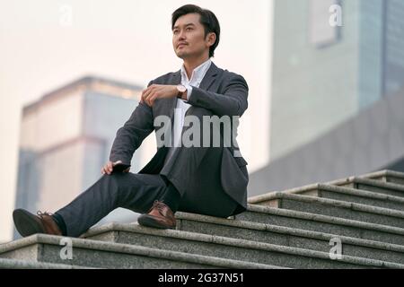 Außenportrait eines erfolgreichen asiatischen Geschäftsmanns, der in Vollanzug auf einer Treppe sitzt Stockfoto