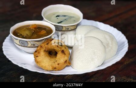 Südindisches Frühstücksmenü, Idid Vadai mit Kokosnuss-Chutney und Sambar, serviert auf einem weißen Keramikteller auf einem Holztisch. Stockfoto